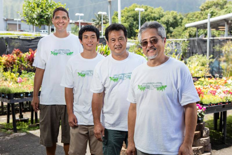 Four smiling men in white, Koolau Farmer t-shirts standing outdoors in front of tables of potted plants